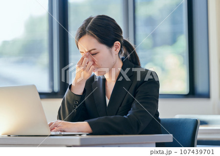 パソコンを操作しながら眉間に手を当てる若い女性 107439701