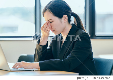 パソコンを操作しながら眉間に手を当てる若い女性 107439702