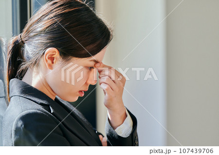 窓際に立ち眉間に手を当てる若い女性 窓際に立ち眉間に手を当てる若い女性 107439706
