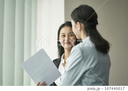 two young asian business women standing by window having a discussion in office two young asian business women standing by window having a discussion in office 107440017