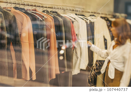 Young woman looking new clothes in fashion store in boutique, shopping concept. Blurred view through glass Young woman looking new clothes in fashion store in boutique, shopping concept. Blurred view through glass 107440737