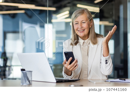 Happy senior business woman sitting in the office at the desk and looking at the phone screen, got good news and message. 107441160
