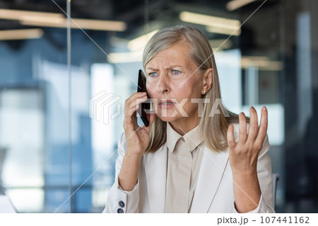 Close-up photo. Worried and upset senior business woman talking on the phone in the office and spreading her hands. 107441162