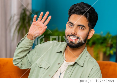 Indian man smiling friendly at camera, waving hands gesturing hello, hi, greeting at home on sofa Indian man smiling friendly at camera, waving hands gesturing hello, hi, greeting at home on sofa 107441434