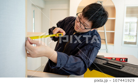 Asian male furniture assembler using tape measure 107441929