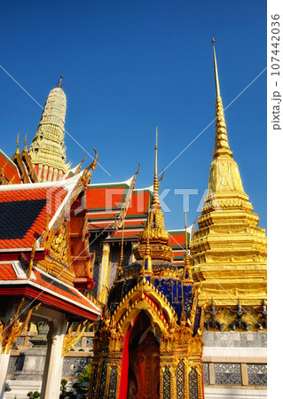 An enchanting low-angle shot frames a Thai temple's red rooftops, opulent golden highlights, and an imposing white spire against the canvas of a crystal-clear blue sky. 107442036