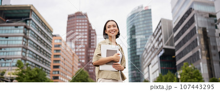 Portrait of young asian woman, looking happy and confident, going to work or university, city skyscrappers behind her, holding laptop and notebook Portrait of young asian woman, looking happy and confident, going to work or university, city skyscrappers behind her, holding laptop and notebook 107443295