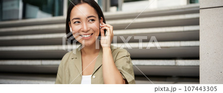 Close up portrait, street photo of asian woman listening music in wireless headphones, smiling, sitting on stairs 107443305