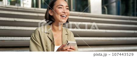 Beautiful asian girl sits on stairs with smartphone. Young korean woman resting outdoors, using mobile phone application Beautiful asian girl sits on stairs with smartphone. Young korean woman resting outdoors, using mobile phone application 107443529