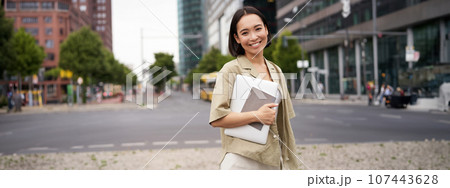 Portrait of young asian woman, looking happy and confident, going to work or university, city skyscrappers behind her, holding laptop and notebook Portrait of young asian woman, looking happy and confident, going to work or university, city skyscrappers behind her, holding laptop and notebook 107443628