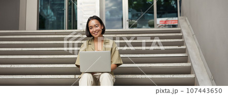 Young asian woman using laptop outdoors. Cute girl student with computer, does her homework, types, sits on stairs outside campus Young asian woman using laptop outdoors. Cute girl student with computer, does her homework, types, sits on stairs outside campus 107443650