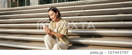 Beautiful asian girl sitting on stairs outside building, using mobile phone, looking at smartphone app and smiling 107443797