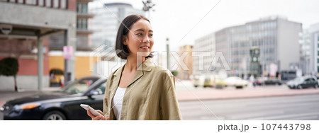 Young woman walking down the city street, turn behind, using map app navigator on smartphone, holding mobile phone 107443798