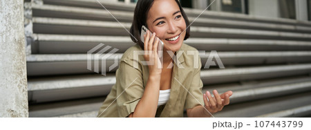 Close up portrait of smiling asian girl talks on mobile phone, sits outside on street stairs. Young woman calling friend on smartphone 107443799