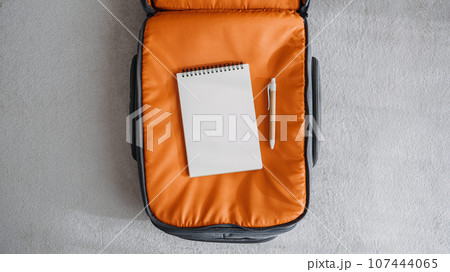 Travel Packing List For Vacation mockup. Travel Packing Checklist For Holiday Or Business Trip. Open empty notepad with pen in orange suitcase on bed at home 107444065