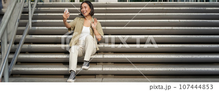 Portrait of asian beautiful girl takes selfie on smartphone. Young korean woman sits on stairs outdoors and makes photos on mobile phone 107444853