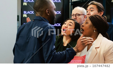 Crazy people breaking store line and pushing security guard to enter clothing retail shop on black friday. Aggressive crowd behind red tape at fashion boutique, big sales. Handheld shot. 107447482