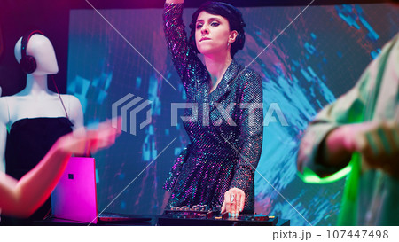 Female DJ mixing music at club on stage, partying with group of funky people in discotheque. Young woman having fun jumping with persons on dance floor, using audio panel station. 107447498