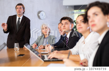 Displeased male manager scolding at young, adult, mature office workers sitting with reports and laptops at table in meeting room 107449739