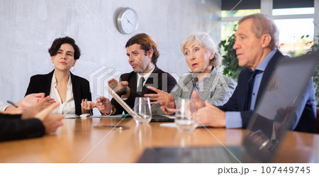 Office coworkers of different ages discussing topic of day while sitting at table with laptop and notes 107449745