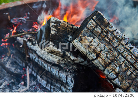 Close up of ashen logs smoking with embers and orange and yellow flames in background asset 107452200