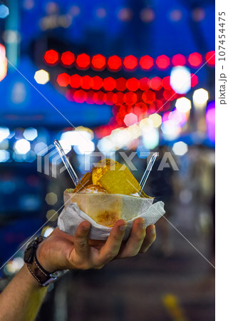 Hand holding coconut and mango ice cream in coconut shell against lights of Jalan Alor street food market in Kuala Lumpur 107454475