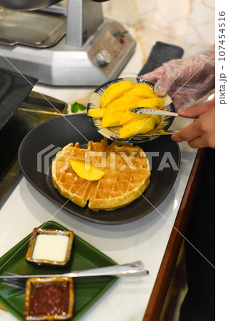 the cook putting fresh sliced mango fruit on baked belgian waffles with tongs 107454516