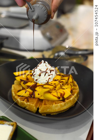 the cook putting chocolate syrup on fresh sliced mango fruit on baked belgian waffles the cook putting chocolate syrup on fresh sliced mango fruit on baked belgian waffles 107454524