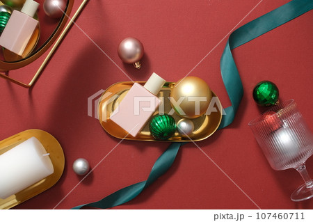 Scene for advertising and branding product with Christmas concept. A pink cosmetic bottle placed on golden tray with decorations: baubles, ribbons and candles on red background Scene for advertising and branding product with Christmas concept. A pink cosmetic bottle placed on golden tray with decorations: baubles, ribbons and candles on red background 107460711
