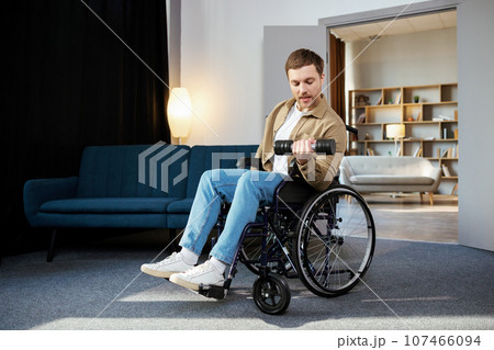 Physical rehabilitation for disabled people. Happy guy in wheelchair making exercises with dumbbell at home. Cheerful impaired young man working out with weights indoors Physical rehabilitation for disabled people. Happy guy in wheelchair making exercises with dumbbell at home. Cheerful impaired young man working out with weights indoors 107466094
