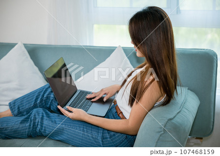 Asian young woman smiling sitting relaxing on sofa using laptop in living room at home, freelance working on computer project, Happy female typing email on notebook computer 107468159