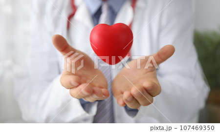 Doctor cardiologist holds hands and airborne red toy heart closeup Doctor cardiologist holds hands and airborne red toy heart closeup 107469350