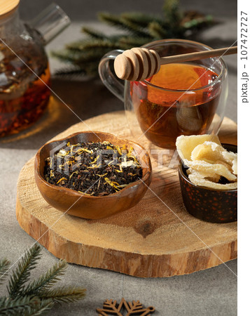 Glass cup with tea and wooden spoon for honey. On a round wooden tray with candied ginger and tea leaves in a bowl. Close-up Glass cup with tea and wooden spoon for honey. On a round wooden tray with candied ginger and tea leaves in a bowl. Close-up 107472727