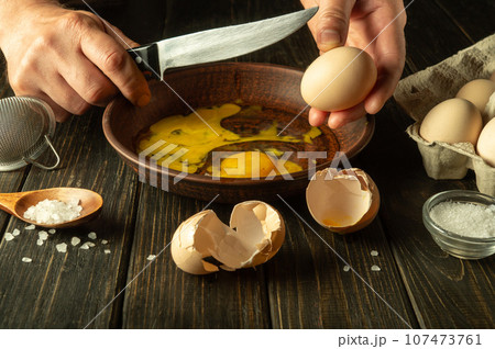 The cook prepares eggs on the kitchen table for an omelette. The chef hands hold a knife to break an egg. Healthy egg diet 107473761