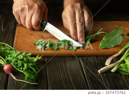 Cooking a dietary salad for lunch from fresh vegetables and sorrel leaves. The chef hands cut sorrels on a kitchen board with a knife. Fresh vegetable useful diet. 107473762
