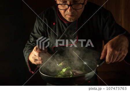 Senior cook or chef prepares vegetables in a frying pan. Cooking healthy vegetarian food and dishes on a dark background. The concept of cooking vegan food. Senior cook or chef prepares vegetables in a frying pan. Cooking healthy vegetarian food and dishes on a dark background. The concept of cooking vegan food. 107473763