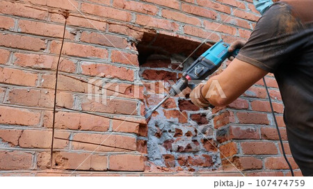 a builder in a black T-shirt and gloves chiseling the brickwork of a house wall with a pneumatic drill, working to destroy a brick wall to make a window opening, drilling the external wall 107474759