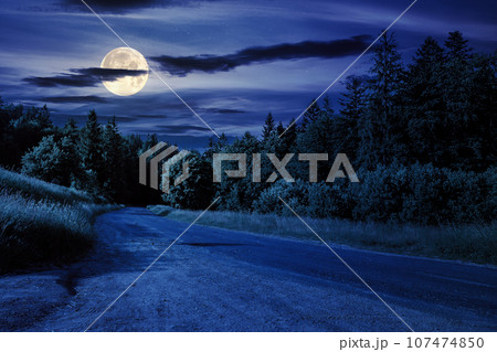 asphalt road with cracks winding through shaded coniferous forest at night. beautiful countryside scenery in full moon light 107474850