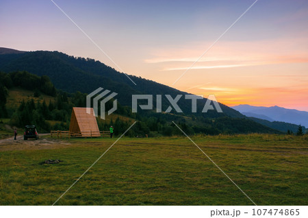 sunset in carpathian mountains. distant rural valley with rolling hills between forested slopes in hazy atmosphere. wooden shelter on the grassy meadow 107474865