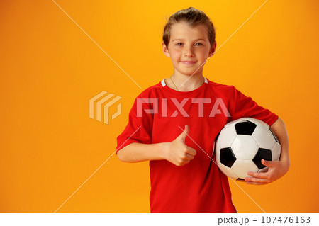 Teen boy soccer player with football ball against yellow background Teen boy soccer player with football ball against yellow background 107476163