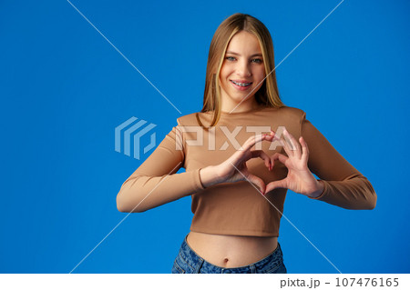 Friendly teen girl shows heart love gesture on blue background Friendly teen girl shows heart love gesture on blue background 107476165