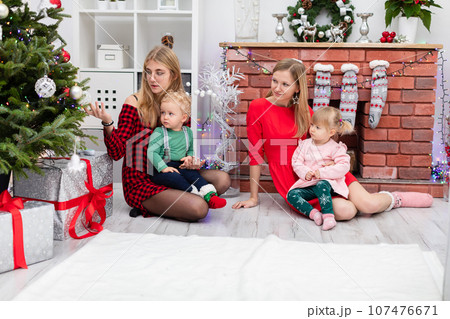 Two women sit together with their young children by the Christmas tree Two women sit together with their young children by the Christmas tree 107476671