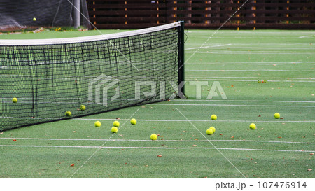 Tennis court, yellow tennis balls are scattered on the green field near the net. Active recreation, sports competitions, equipment for games. Tennis court, yellow tennis balls are scattered on the green field near the net. Active recreation, sports competitions, equipment for games. 107476914