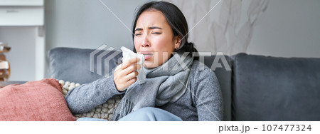 Close up of korean girl sneezing in napking, sitting at home in scarf, feeling unwell, concept of winter, heating cost and people Close up of korean girl sneezing in napking, sitting at home in scarf, feeling unwell, concept of winter, heating cost and people 107477324