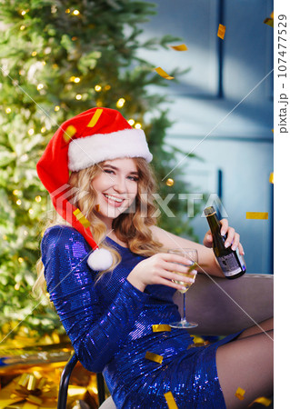 woman in a shiny blue dress in santa's hat with champagne by the Christmas tree. 107477529