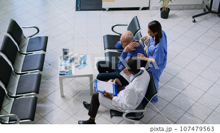 Asian nurse helping senior patient to take off cervical neck collar in hospital reception, african american doctor explaining medical expertise. Elderly man having checkup visit consultation 107479563
