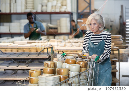 Elderly worker in ceramics factory carries many different clay ceramic cups and plates in cart 107481580