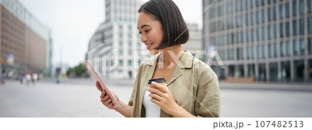 Portrait of smiling korean girl looks at tablet, reads on street, drinks takeaway coffee Portrait of smiling korean girl looks at tablet, reads on street, drinks takeaway coffee 107482513