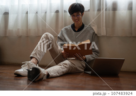 A happy young Asian man enjoys reading a book while sitting on the floor in a room. A happy young Asian man enjoys reading a book while sitting on the floor in a room. 107483924