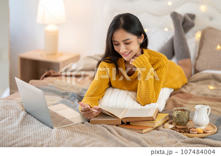 An attractive Asian woman in a cosy knitted sweater is reading a book on her bed, relaxing at home. An attractive Asian woman in a cosy knitted sweater is reading a book on her bed, relaxing at home. 107484004